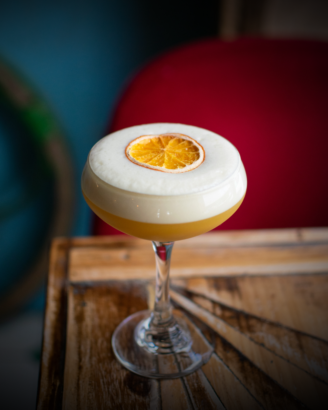 A glass of cocktail garnished with a premium dehydrated orange slice on top, set on a wooden table with a red chair and blue wall in the background.