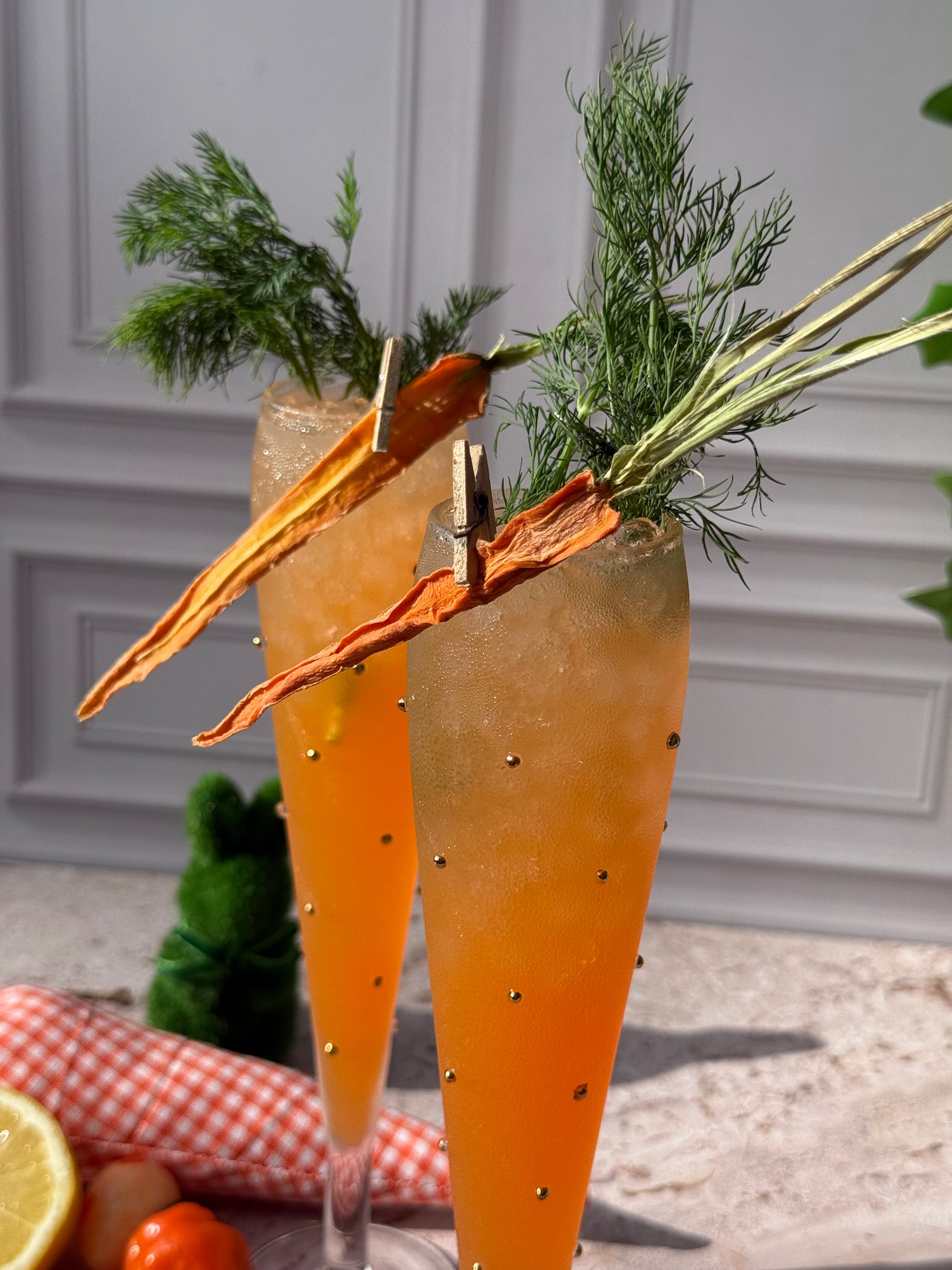 Carrot-shaped cocktails with garnishes on a checkered cloth against a neutral background