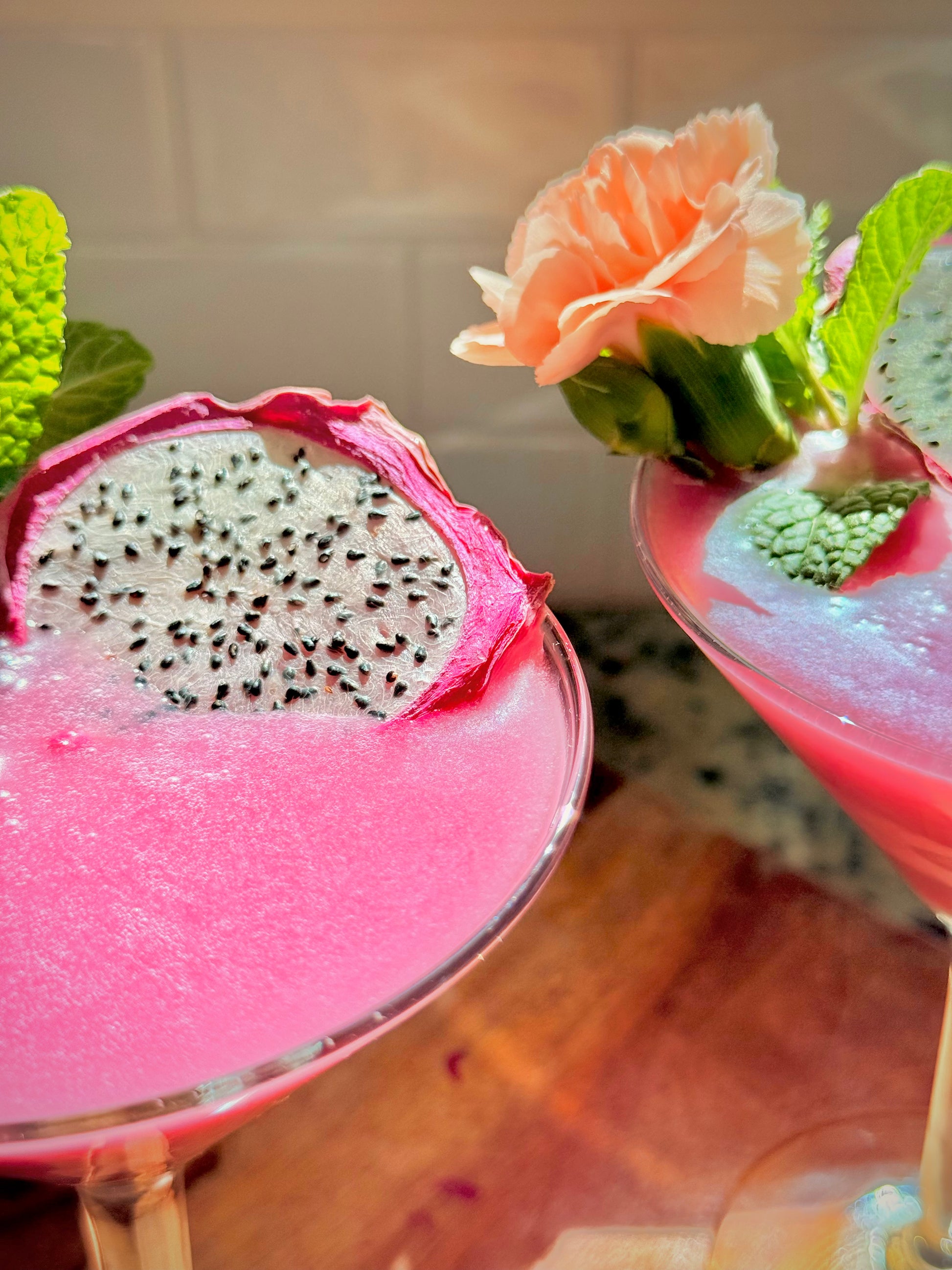 Two pink cocktails with dragon fruit and flowers on a wooden table.
