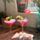 Two pink cocktails with dragon fruit garnishes on a wooden surface, with dragon fruit slices and a vase in the background.