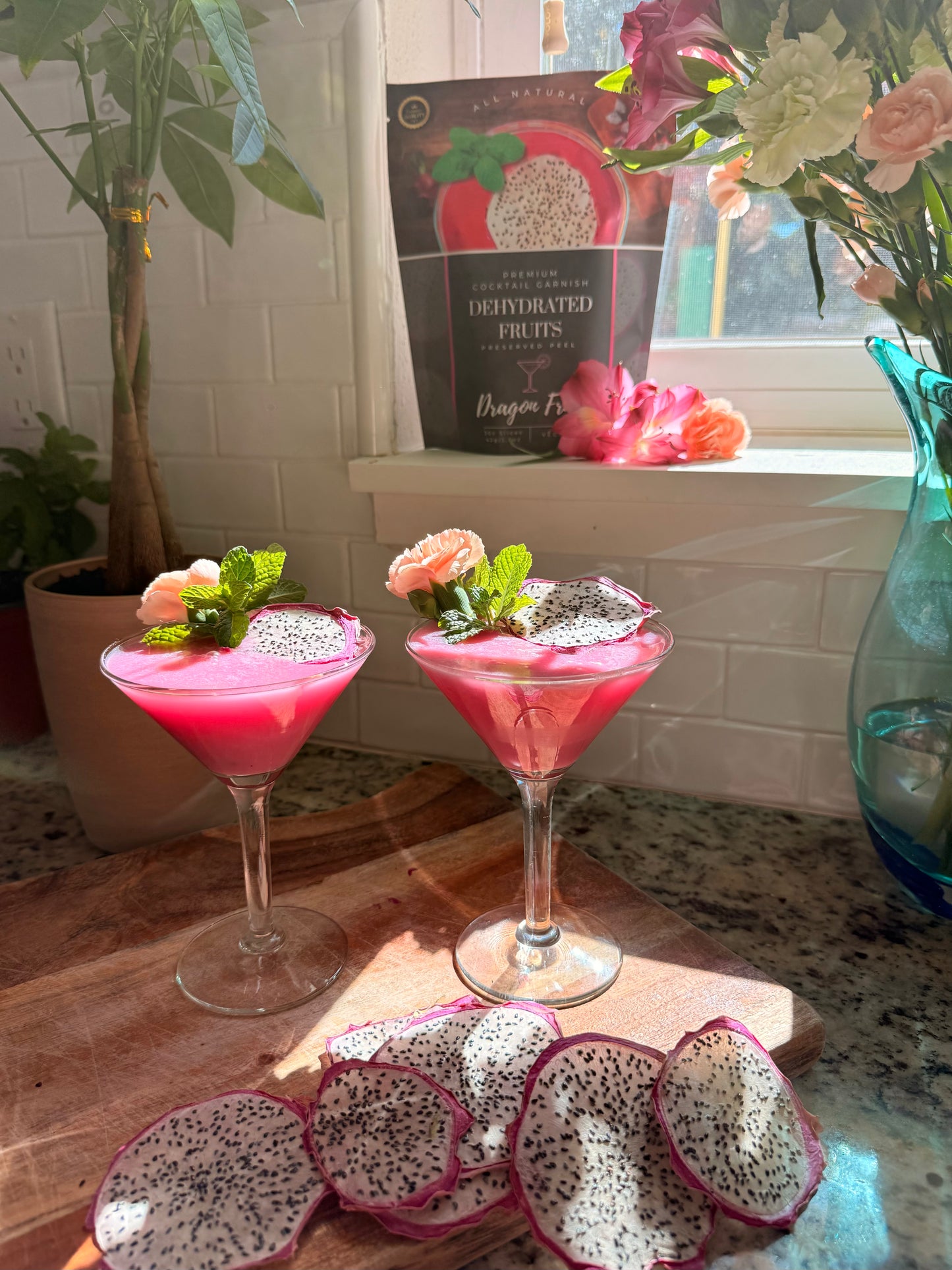 Two pink cocktails with dragon fruit garnishes on a wooden surface, with dragon fruit slices and a vase in the background.