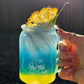 Hand holding a blue and yellow layered drink in a Mason jar with a pineapple wedge on a dark background