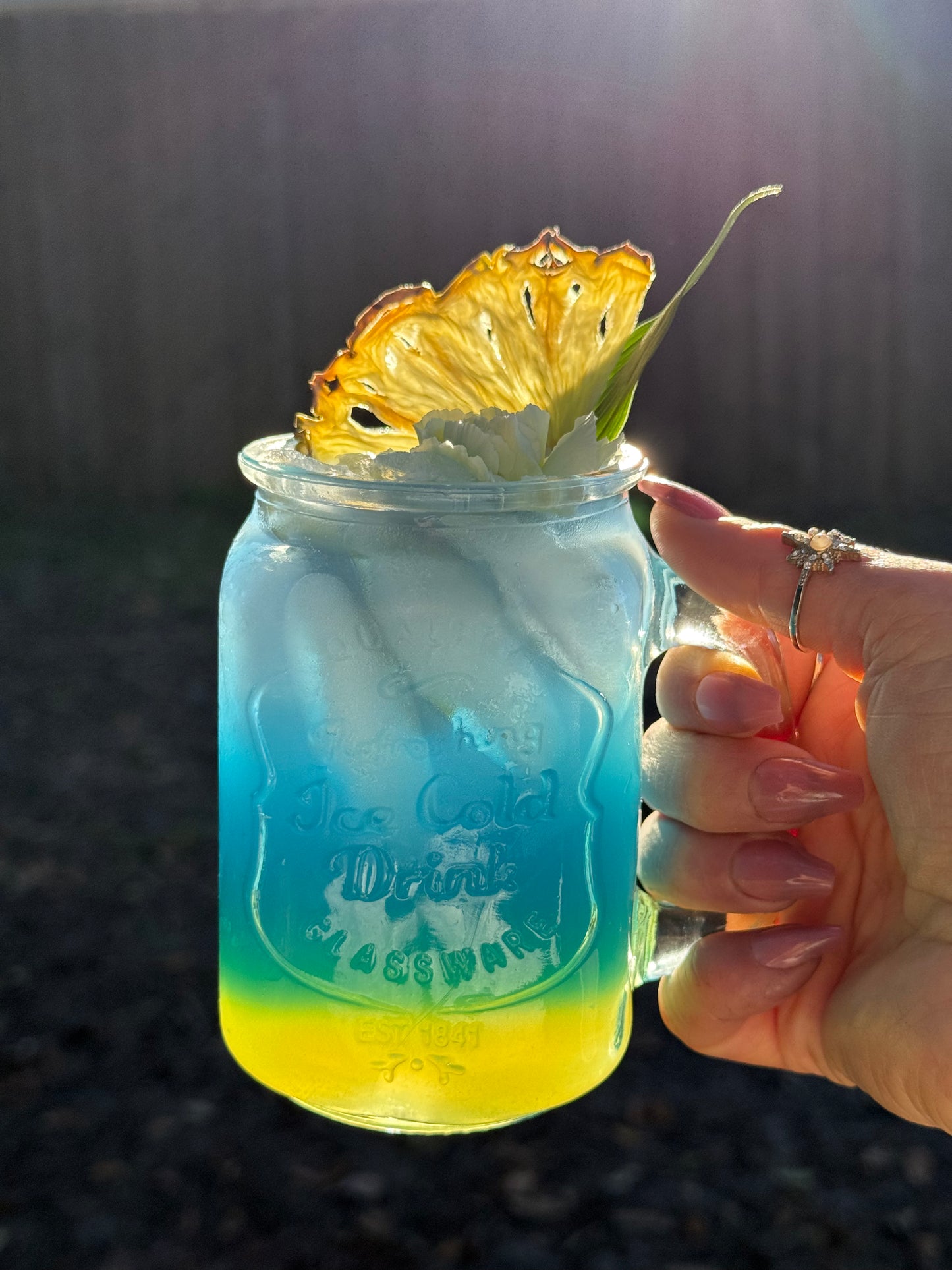 Hand holding a blue and yellow layered drink in a Mason jar with a pineapple wedge on a dark background